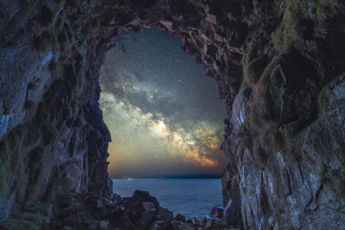 The milky way blazes across the sky, as seen through the entrance of Mousehole Cave.